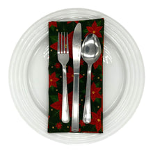 Load image into Gallery viewer, A folded cotton napkin with red poinsettias on a green background, as a place-setting with a plate and cutlery.