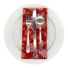 Load image into Gallery viewer, A red dinner napkin with a white, gold, and red snowflake pattern, used as a place-setting with plate and cutlery.