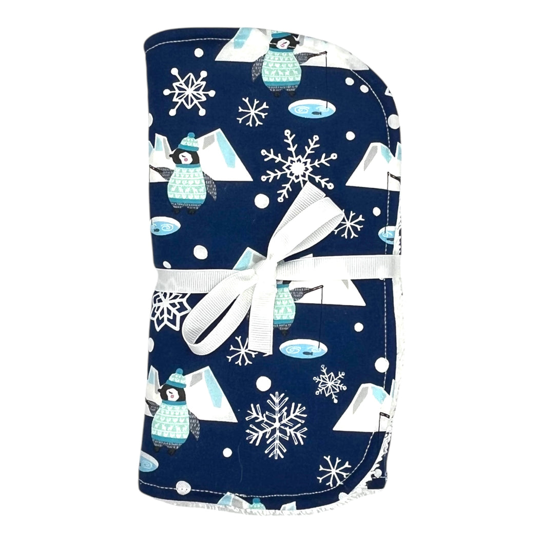 Folded burpcloth tied with white ribbon. Design features penguins fishing, icebergs, and snowflakes.