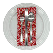 Load image into Gallery viewer, Red napkin with white Christmas-themed pattern. Shown as place setting on a plate with cutlery.
